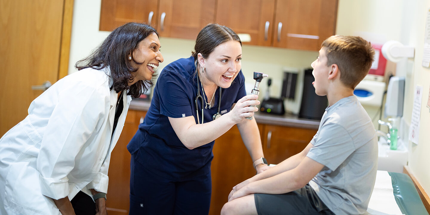 A medical student uses an otoscope to examine a child’s throat in a medical exam room while another clinician observes.