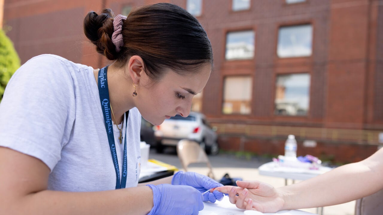 A female medical student collects a blood sample from a patient's finger at an outdoor clinic table.